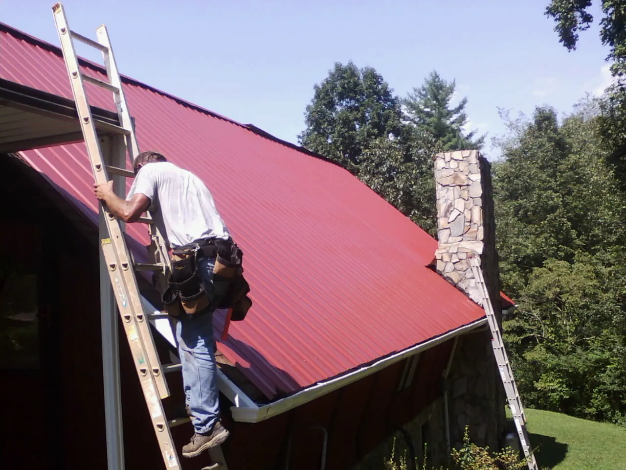 Roofer on a ladder beside a red metal roof and stone chimney — Soffit & Fascia Repair by Roofing Brothers in Manchester
